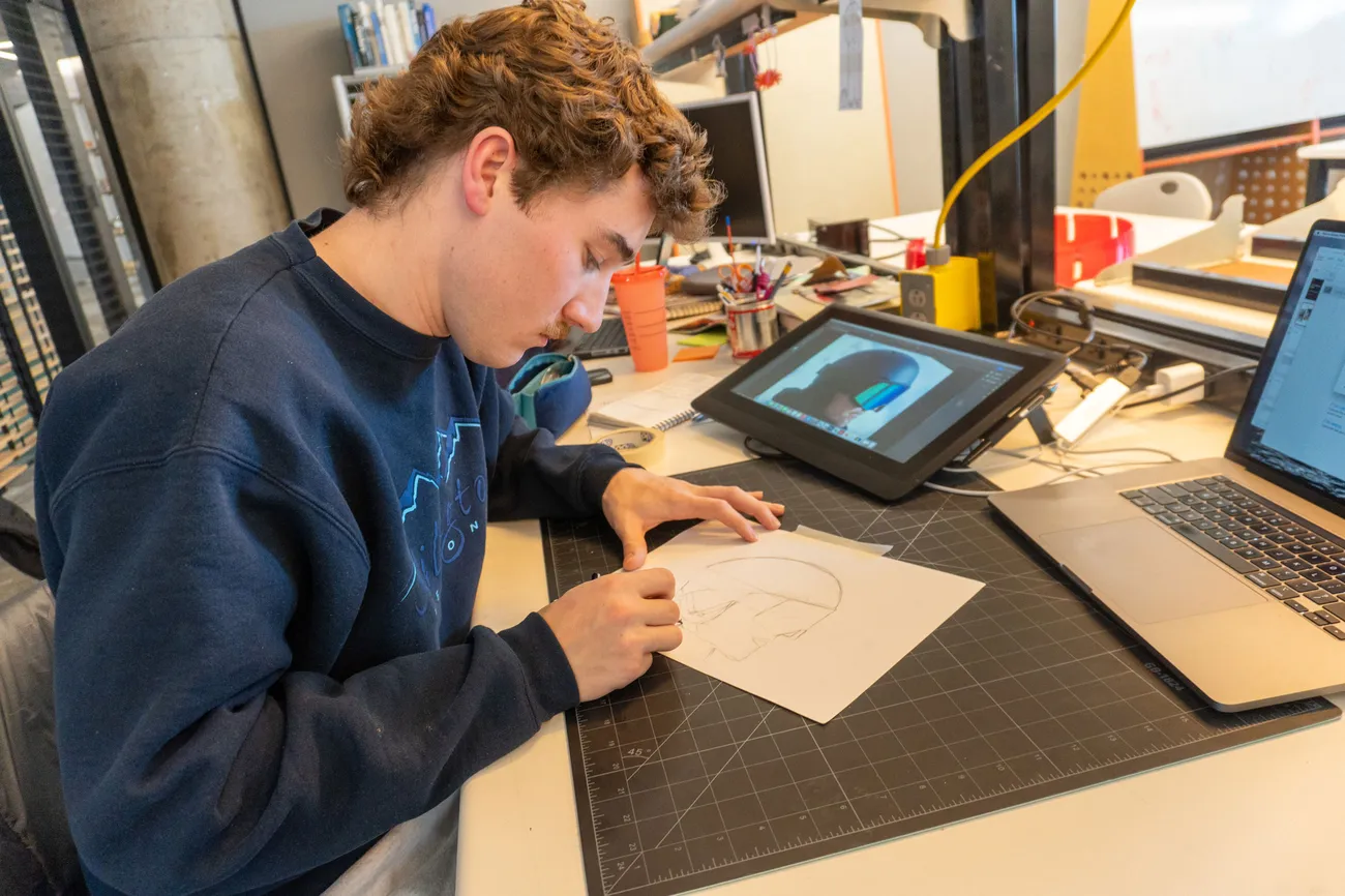 A student sits at a table working on a drawing. A tablet with an image of a helmet is on the table.