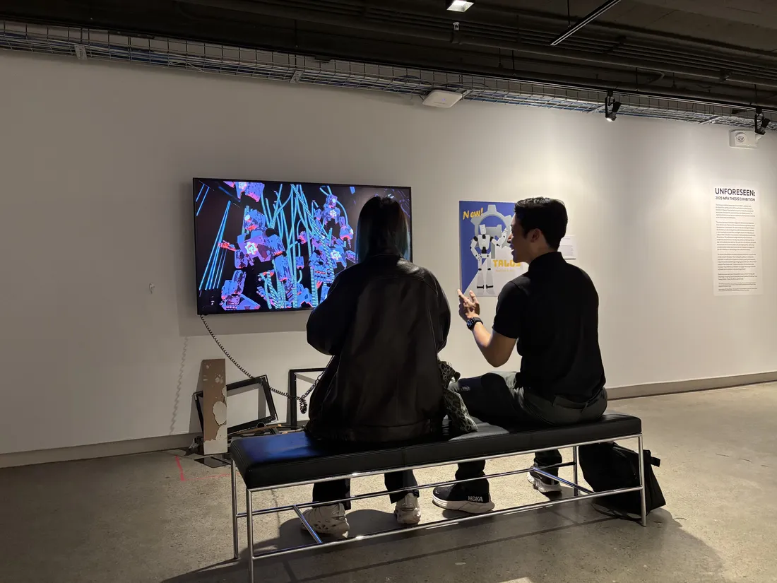 Two people sit on a bench in a gallery looking at a large screen displaying computer-generated work.
