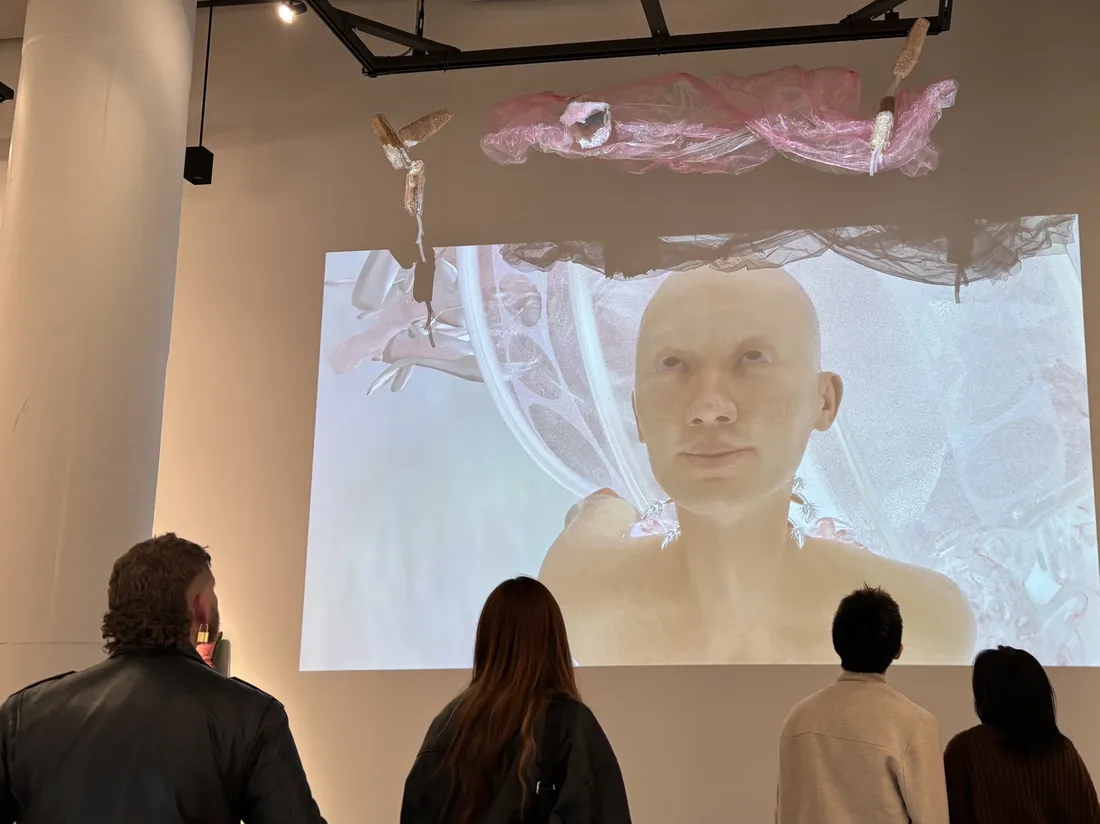 People look at a projection on a gallery wall of a computer-generated image of a bald human being.