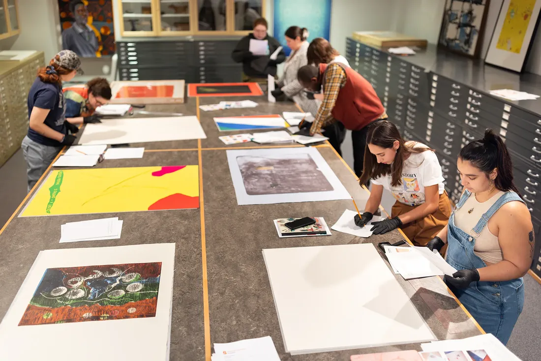 Students stand on either side of a large, long table looking at prints by Helen Frankenthaler.