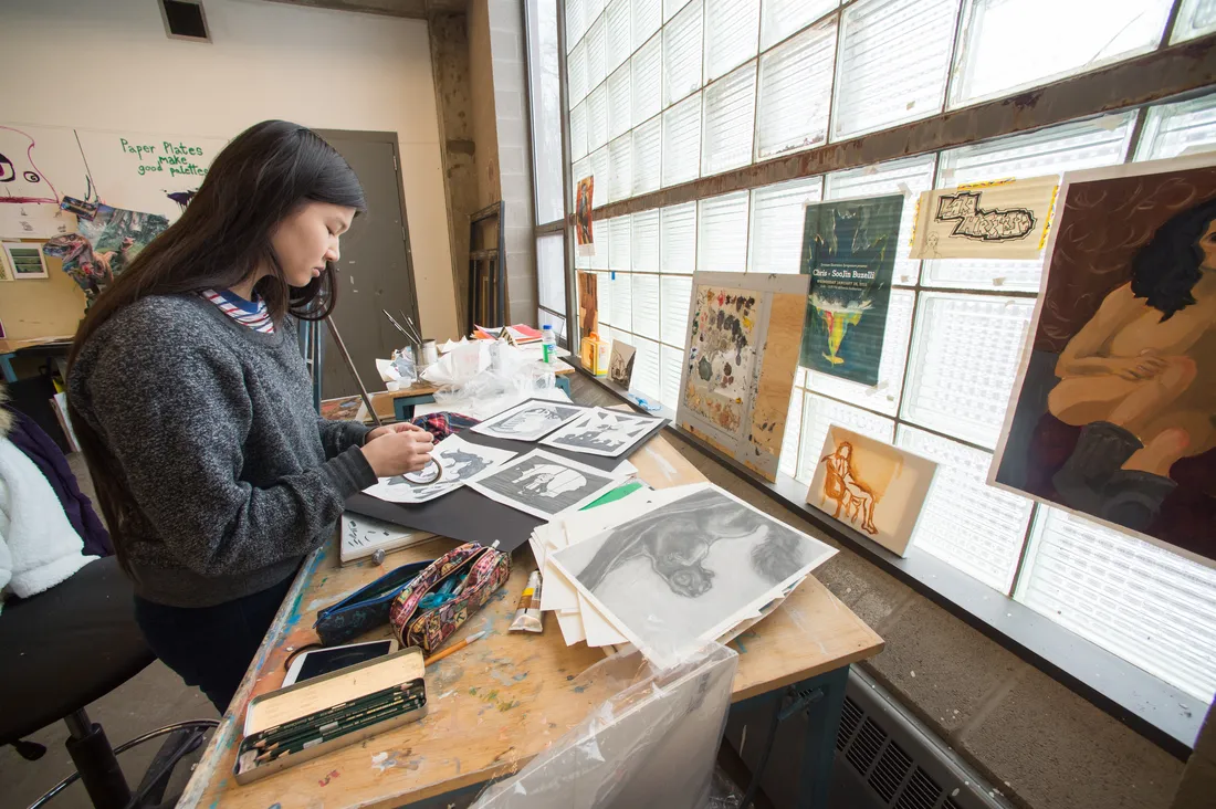 A student stands at a drawing table covered in illustrations. The student is working on an illustration.