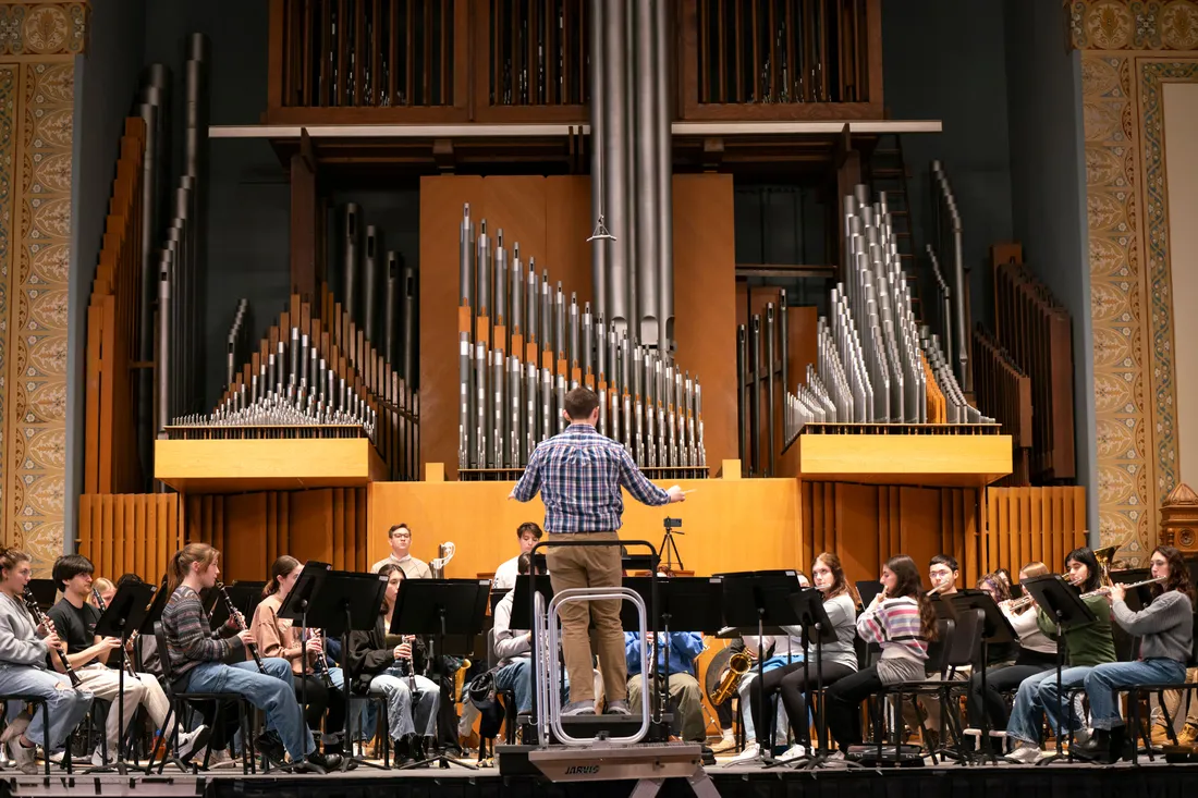 A student conductor with back to the camera stands in front of a wind ensemble.