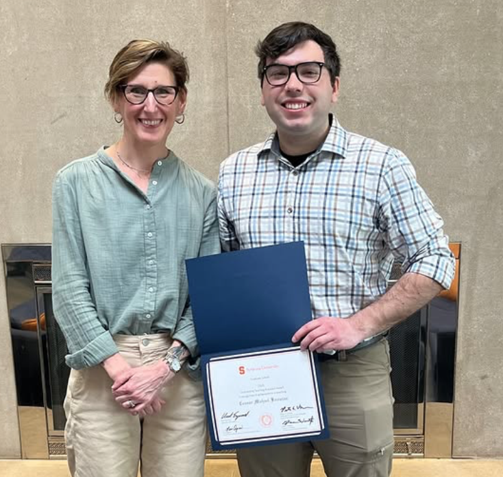 A faculty member and student pose and smile. The student is holding an awards certificate.