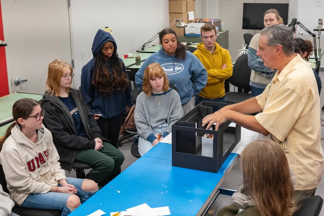 A teacher stands at a table working with a model of a stage while a group of students look on.