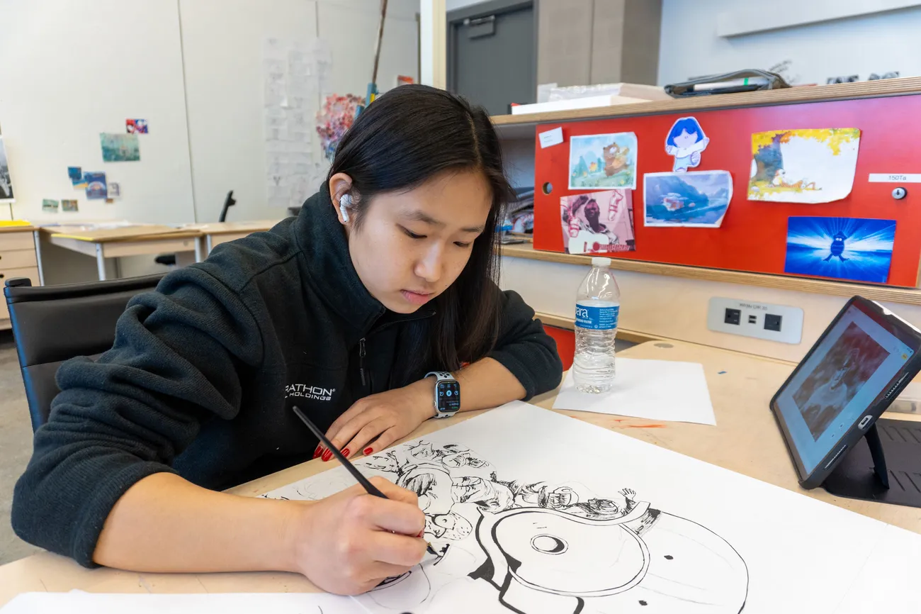 A student sits at a desk and draws on paper.