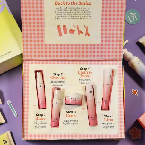 An open pastel pink beauty gift box with a gingham pattern displays a five-step skincare and makeup routine featuring pink cosmetic products for base, cheeks, eyes, lashes and brows, and lips. The box is surrounded by colorful beauty items and decorative accents on a purple background.