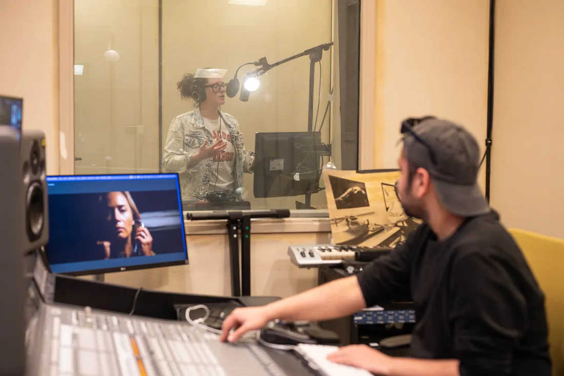 A person sits at a recording console in a recording studio and looks through the window at another person testing a microphone in a sound booth.